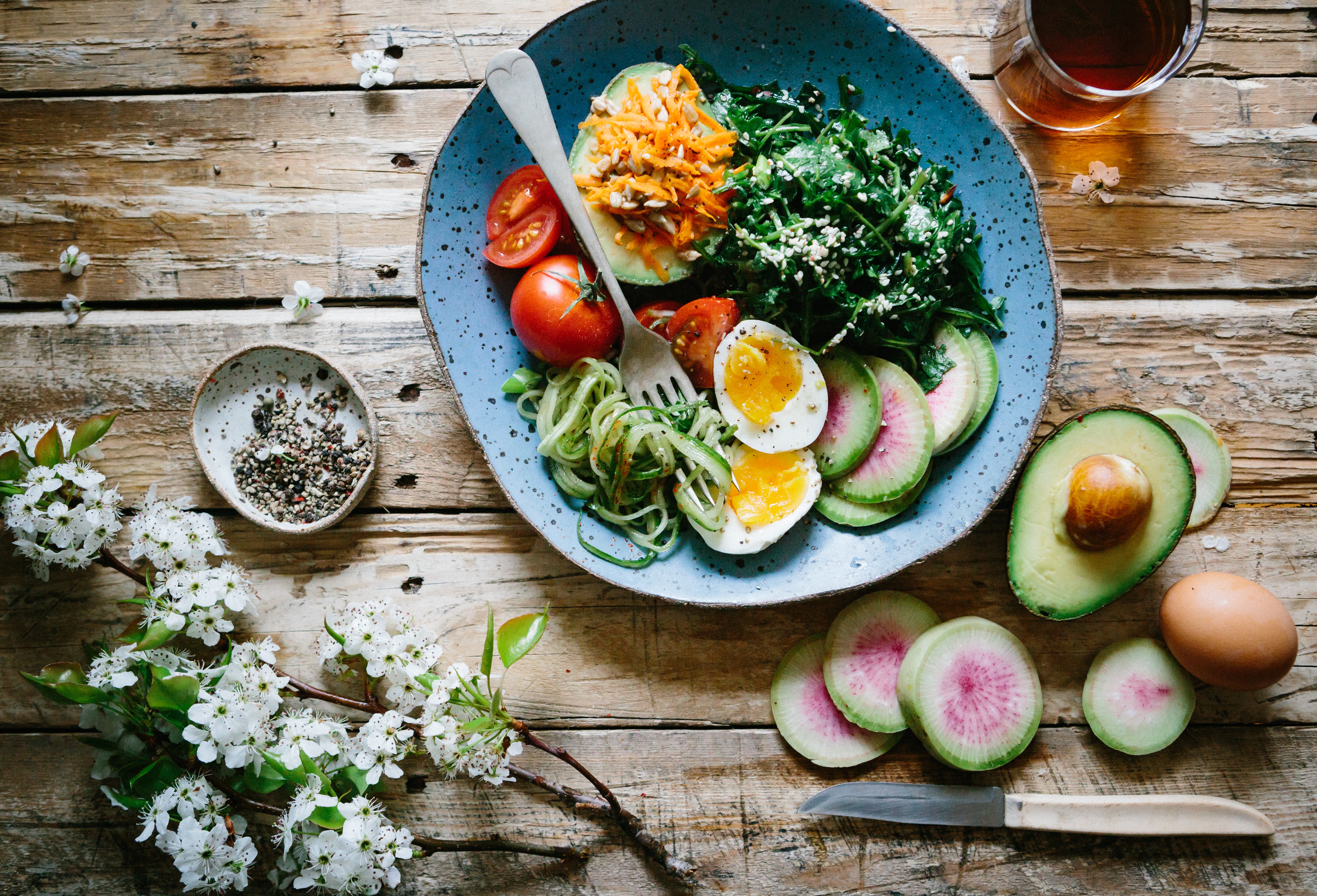 Alt: a salad bowl with healthy foods, surrounded by flowers and tableware. Image courtesy of Unsplash, by Brooke Lark.
